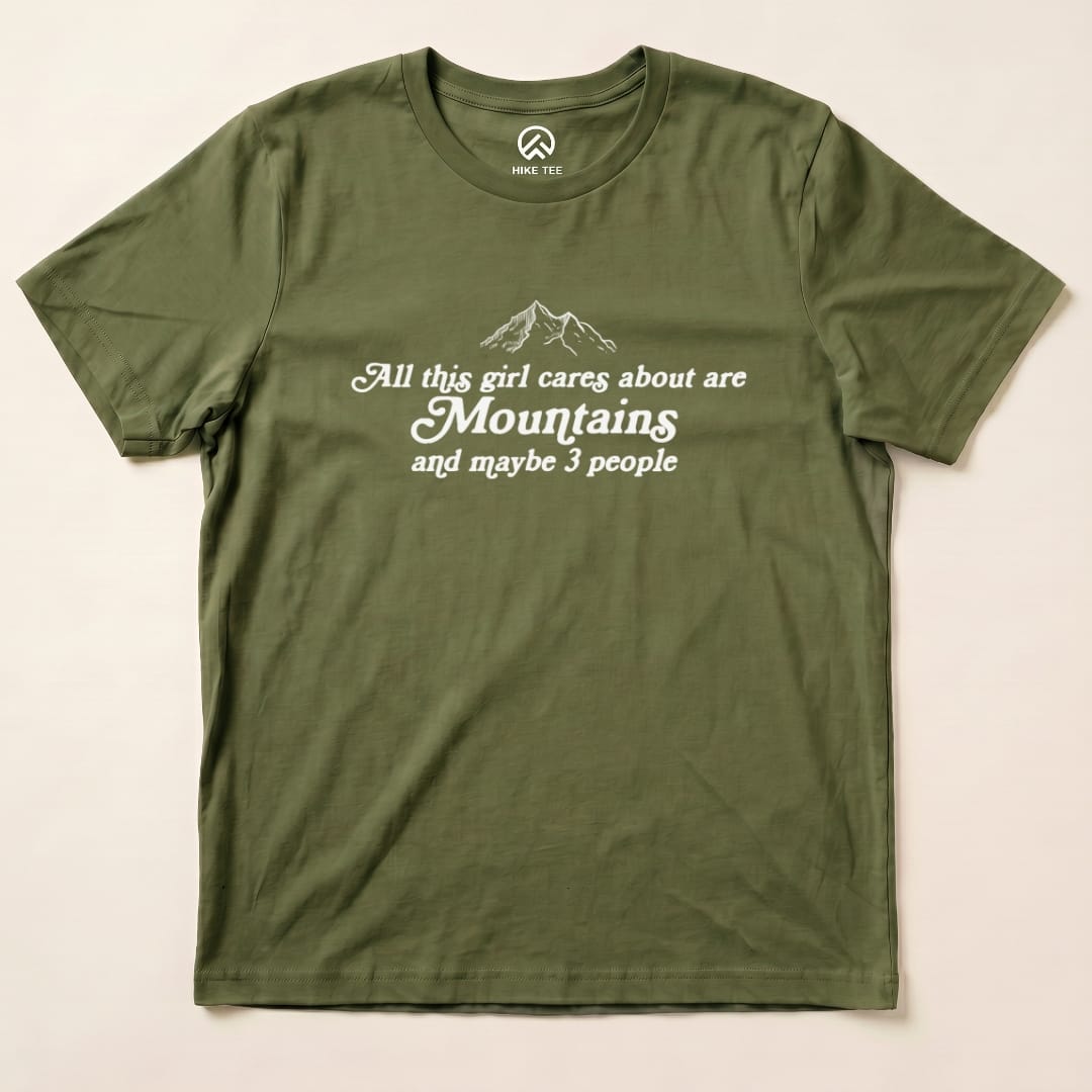 The graphic t-shirt features the slogan "All this girl cares about are Mountains and maybe 3 people" on an olive green background.