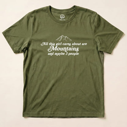 The graphic t-shirt features the slogan "All this girl cares about are Mountains and maybe 3 people" on an olive green background.