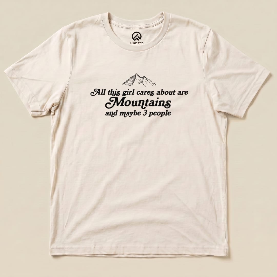 A flat graphic t-shirt featuring the phrase "All this girl cares about are Mountains and maybe 3 people" with mountain illustrations.