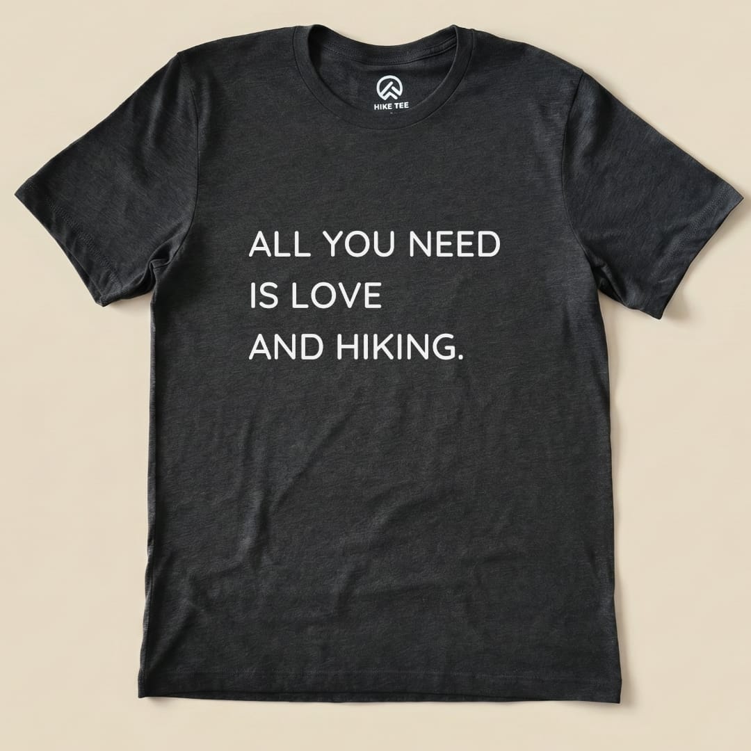 A flat graphic t-shirt in dark gray featuring the slogan "ALL YOU NEED IS LOVE AND HIKING," showcasing the All You Need Hiking Shirt design.