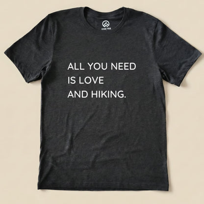 A flat graphic t-shirt in dark gray featuring the slogan "ALL YOU NEED IS LOVE AND HIKING," showcasing the All You Need Hiking Shirt design.