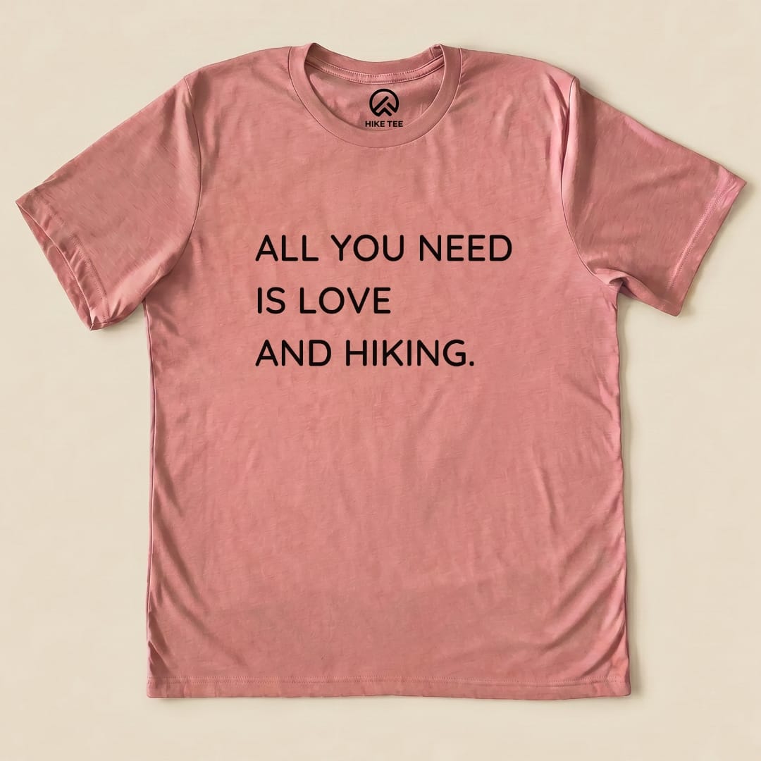 A pink graphic t-shirt featuring the phrase "ALL YOU NEED IS LOVE AND HIKING," labeled as the All You Need Hiking Shirt.