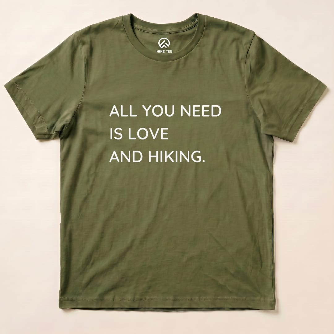 A flat olive green graphic t-shirt displays the text "ALL YOU NEED IS LOVE AND HIKING," showcasing the All You Need Hiking Shirt design.