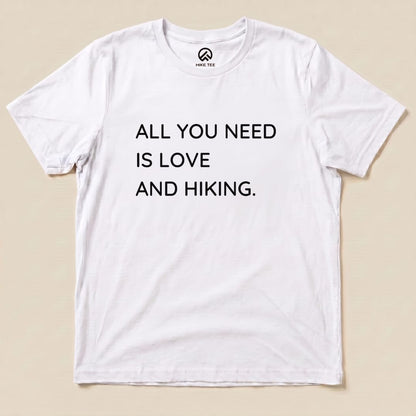 A white graphic t-shirt featuring the slogan "ALL YOU NEED IS LOVE AND HIKING," showcasing a fun hiking theme.