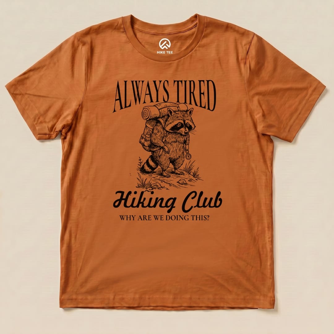 Graphic t-shirt in rust color featuring a raccoon with a backpack, text reads "Always Tired Hiking Club" and "Why Are We Doing This?".