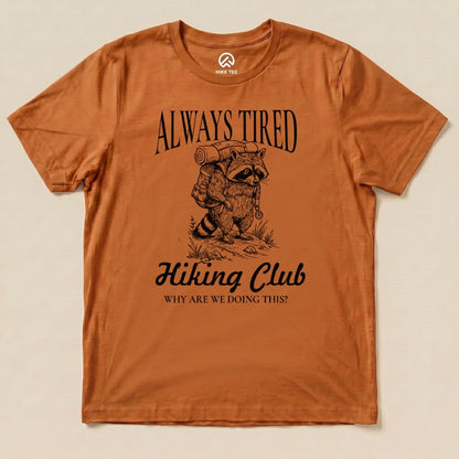 Graphic t-shirt in rust color featuring a raccoon with a backpack, text reads "Always Tired Hiking Club" and "Why Are We Doing This?".