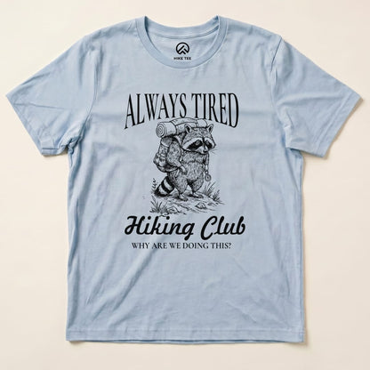 The Always Tired Club Shirt features a raccoon carrying a backpack, with the text "Always Tired Hiking Club" and "Why are we doing this?" printed below.
