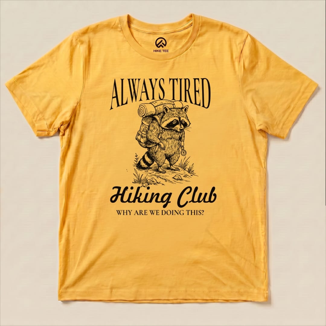 A yellow graphic t-shirt featuring a raccoon with a backpack and the slogan "Always Tired Hiking Club" with the question "WHY ARE WE DOING THIS?