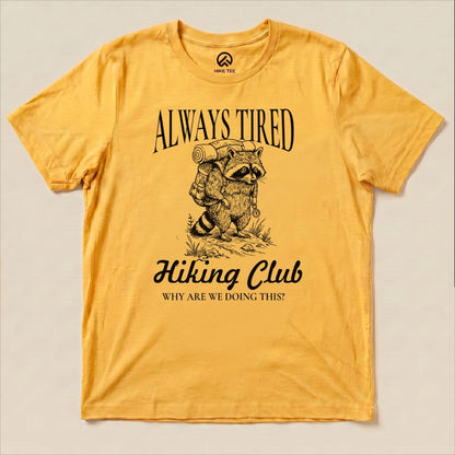 A yellow graphic t-shirt featuring a raccoon with a backpack and the slogan "Always Tired Hiking Club" with the question "WHY ARE WE DOING THIS?
