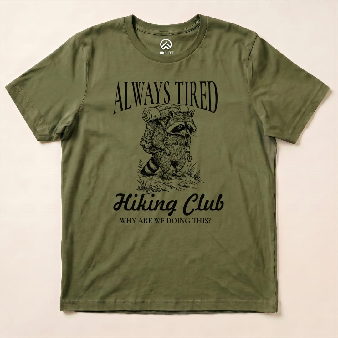 Graphic t-shirt featuring a raccoon with a backpack and the text "Always Tired Hiking Club" above and "WHY ARE WE DOING THIS?" below.