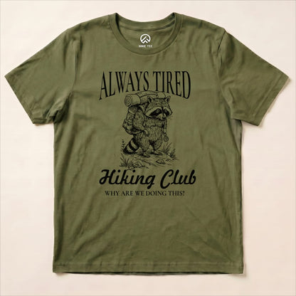 Graphic t-shirt featuring a raccoon with a backpack and the text "Always Tired Hiking Club" above and "WHY ARE WE DOING THIS?" below.