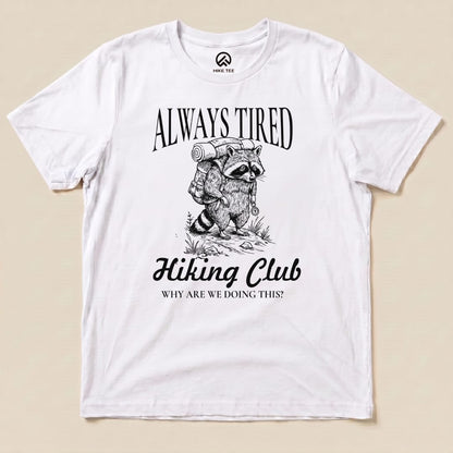 A graphic t-shirt featuring a raccoon carrying a backpack, with the text "Always Tired Hiking Club" and "WHY ARE WE DOING THIS?