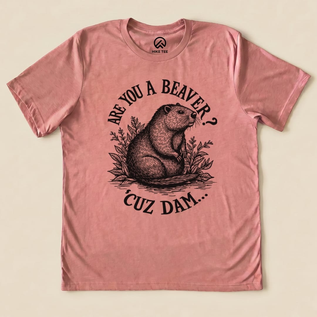A pink graphic t-shirt featuring a beaver surrounded by plants and the text "ARE YOU A BEAVER? 'CUZ DAM..." on the front.