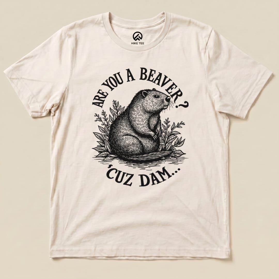 A flat lay image of a cream graphic t-shirt featuring a beaver illustration and the text "ARE YOU A BEAVER? 'CUZ DAM...".