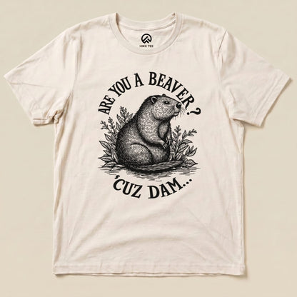 A flat lay image of a cream graphic t-shirt featuring a beaver illustration and the text "ARE YOU A BEAVER? 'CUZ DAM...".