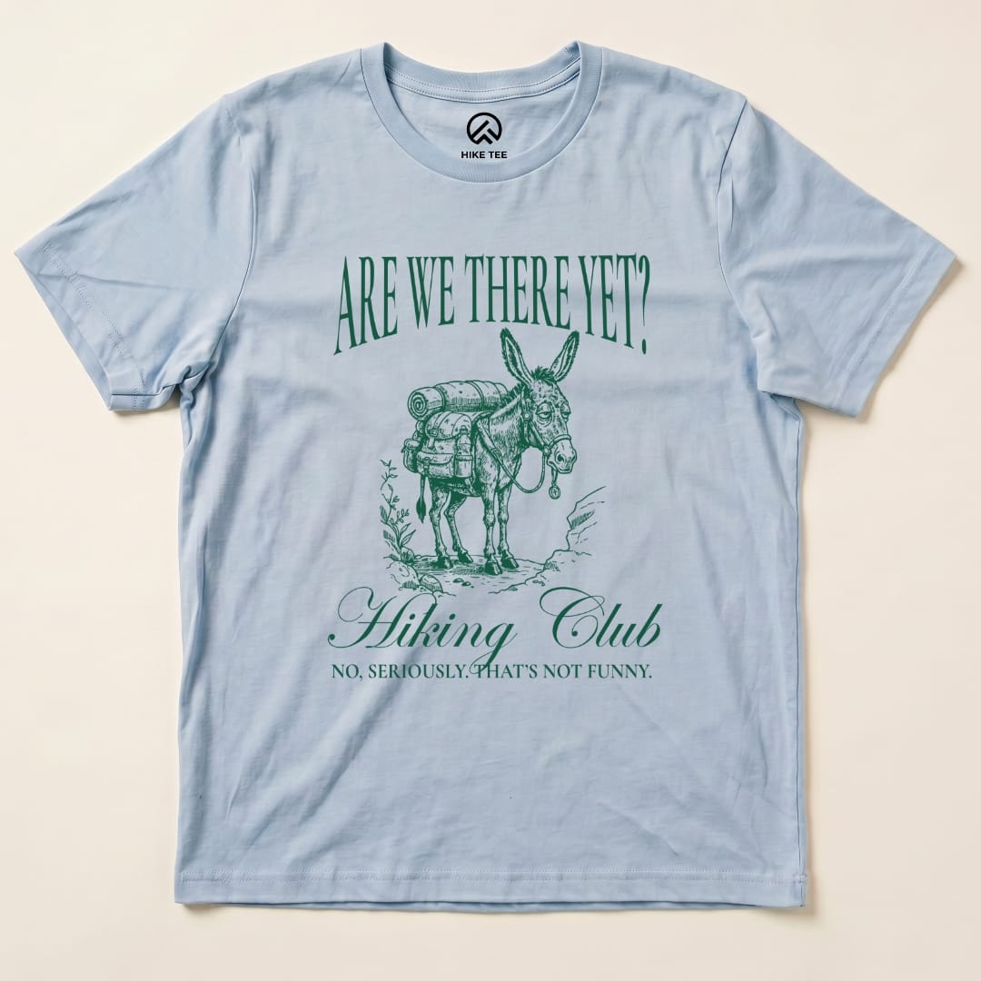 A light blue graphic t-shirt features a donkey with a backpack and the text "Are We There Yet? Hiking Club" above and "NO, SERIOUSLY. THAT'S NOT FUNNY." below, titled "Are We There Donkey Club Shirt.