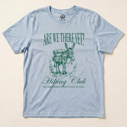 A light blue graphic t-shirt features a donkey with a backpack and the text "Are We There Yet? Hiking Club" above and "NO, SERIOUSLY. THAT'S NOT FUNNY." below, titled "Are We There Donkey Club Shirt.