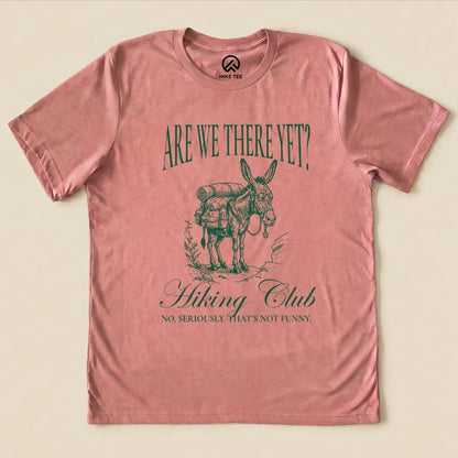 A pink graphic t-shirt featuring a donkey with a backpack, printed with "Are We There Yet? Hiking Club" and the phrase "No, seriously. That’s not funny.