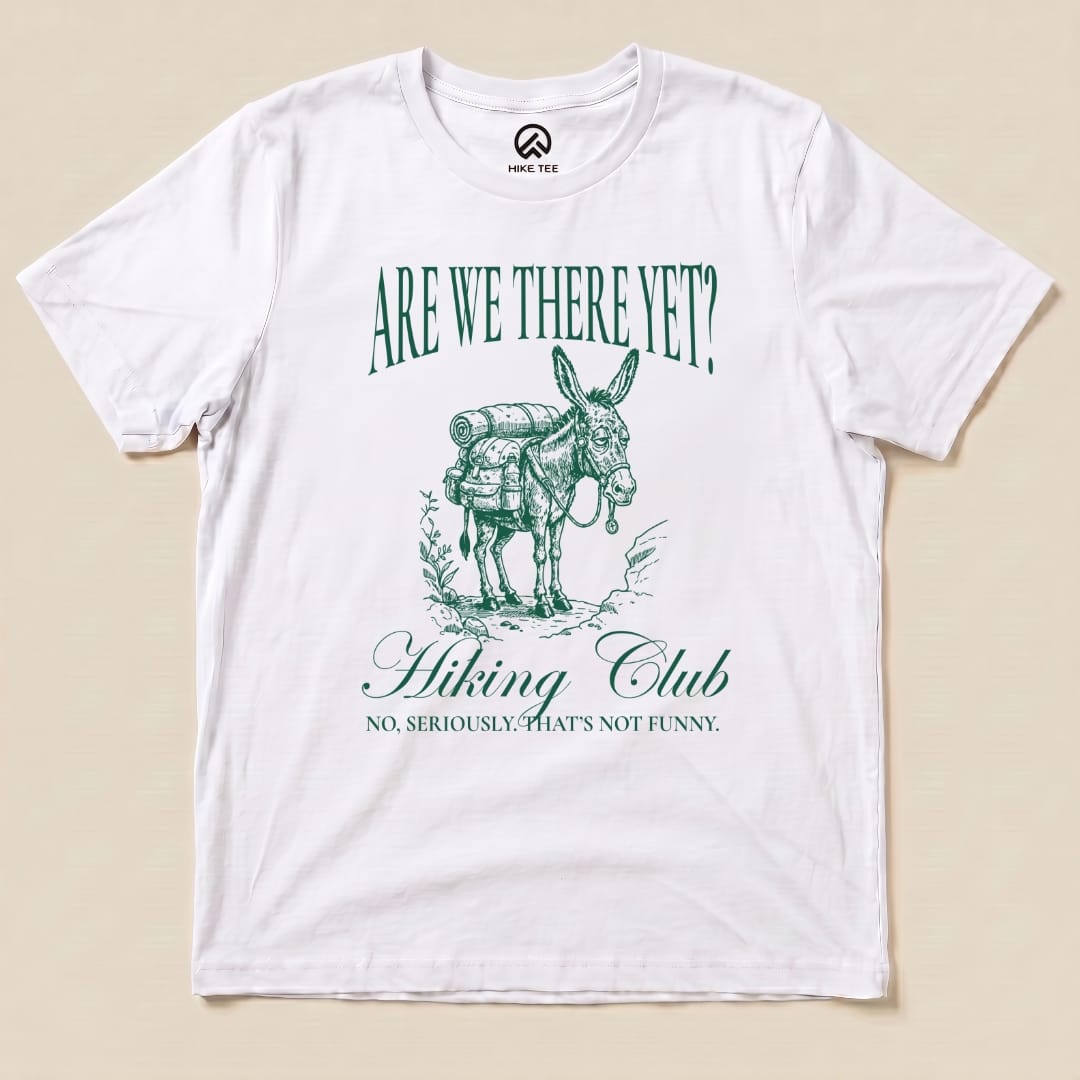 A white graphic t-shirt features a cartoon donkey with a backpack, showcasing the slogan "Are We There Yet? Hiking Club" and "No, seriously. That's not funny.