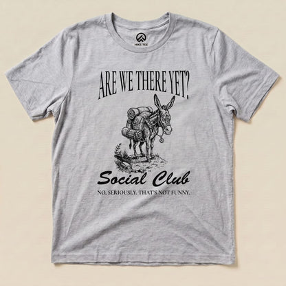 A graphic t-shirt featuring a donkey with a backpack, printed with "Are We There Yet? Social Club" and a humorous tagline below.