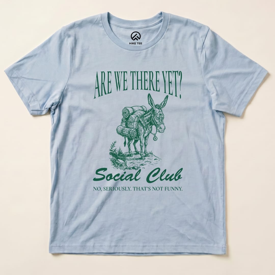 A flat view of a light blue graphic t-shirt featuring a donkey with backpacks and the text "Are We There Yet? Social Club.