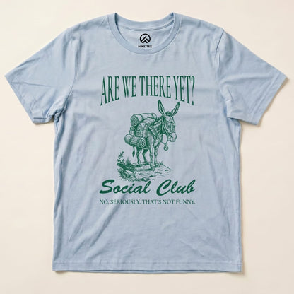A flat view of a light blue graphic t-shirt featuring a donkey with backpacks and the text "Are We There Yet? Social Club.