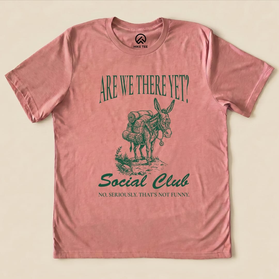 A graphic t-shirt in pink featuring a donkey with backpacks, reading "Are We There Yet? Social Club" and "No, seriously. That's not funny.