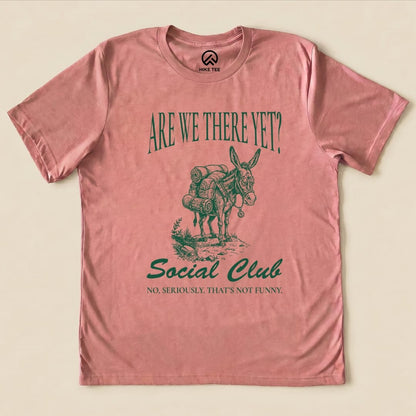 A graphic t-shirt in pink featuring a donkey with backpacks, reading "Are We There Yet? Social Club" and "No, seriously. That's not funny.