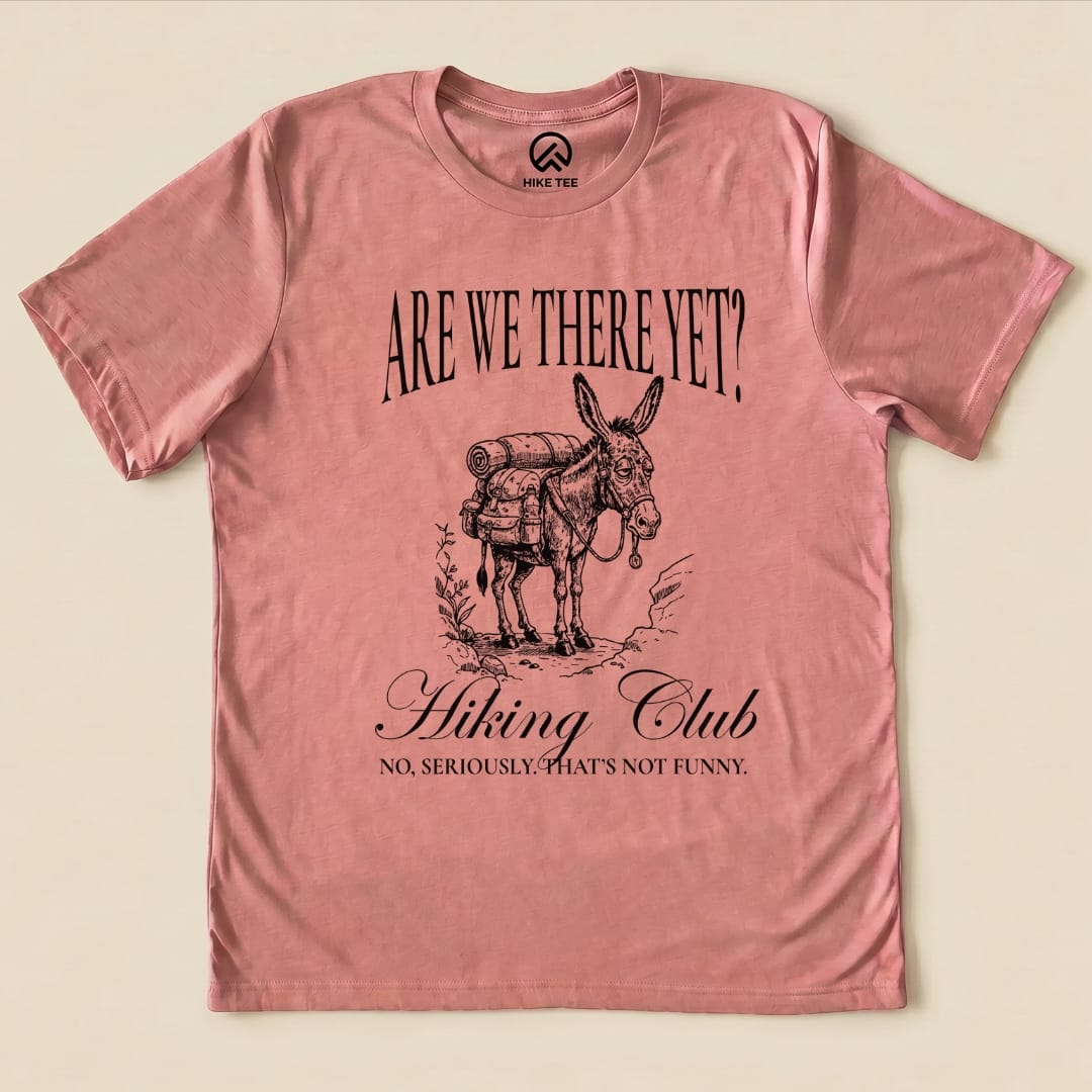 A pink graphic t-shirt featuring the text "Are We There Yet? Hiking Club" above an illustration of a donkey with a backpack.