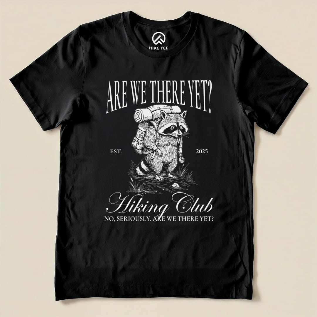 A graphic t-shirt featuring a raccoon carrying a backpack with the text "Are We There Yet Hiking Club" and "No, seriously, are we there yet?
