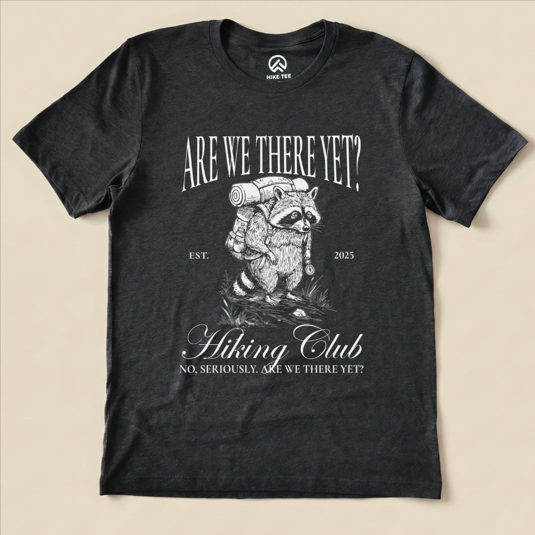 A graphic t-shirt featuring a raccoon carrying a backpack with the slogan "Are We There Yet? Hiking Club" printed above.