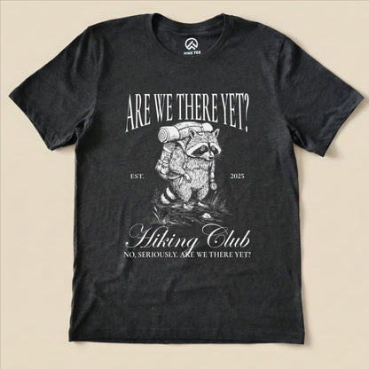 A graphic t-shirt featuring a raccoon carrying a backpack with the slogan "Are We There Yet? Hiking Club" printed above.