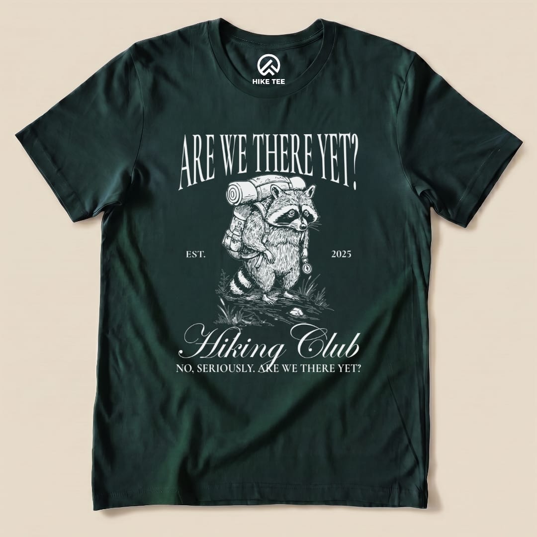 The "Are We There Yet Hiking Club Shirt" features a whimsical raccoon with a backpack, accompanied by playful text and an established date.