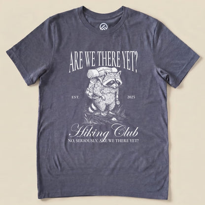Graphic t-shirt featuring a raccoon with a backpack and the slogan "Are We There Yet Hiking Club" above "No, seriously. Are we there yet?