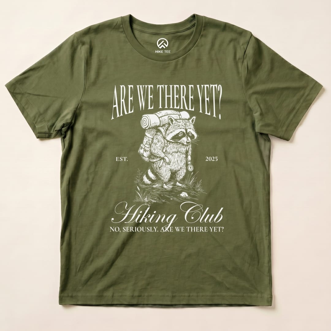 A graphic t-shirt featuring a raccoon with a backpack and the slogan "Are We There Yet Hiking Club" printed above.