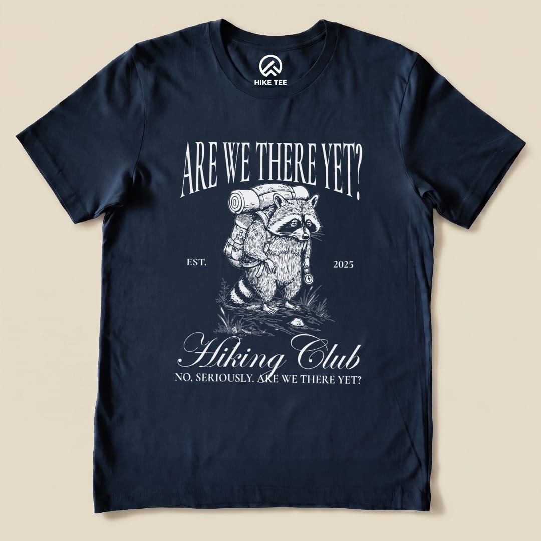 A navy graphic t-shirt features a raccoon with a backpack and the text "Are We There Yet? Hiking Club" above and below in a humorous design.