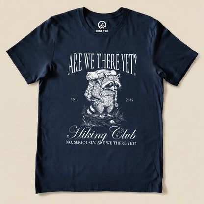 A navy graphic t-shirt features a raccoon with a backpack and the text "Are We There Yet? Hiking Club" above and below in a humorous design.