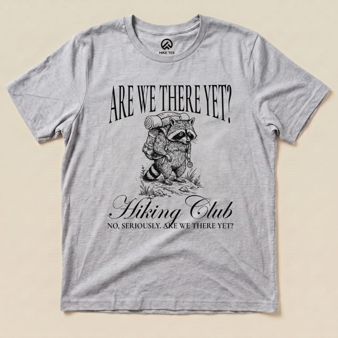 A gray graphic t-shirt featuring a raccoon carrying a backpack, with the text "Are We There Yet? Hiking Club" below.