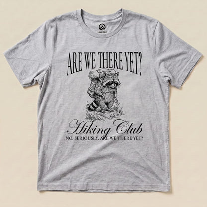 A gray graphic t-shirt featuring a raccoon carrying a backpack, with the text "Are We There Yet? Hiking Club" below.