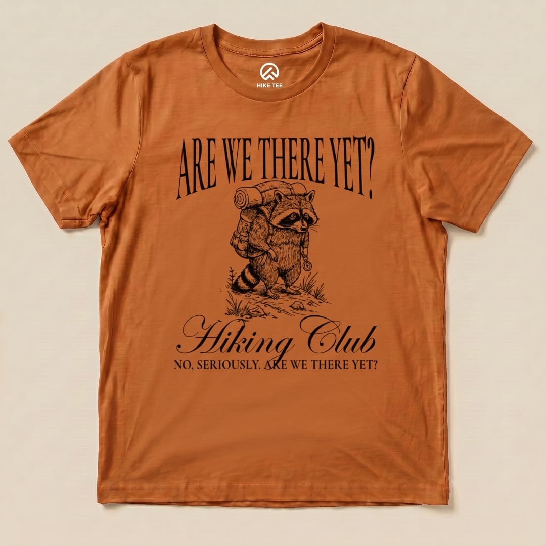 Graphic t-shirt in orange featuring a raccoon with a backpack and the slogan "Are We There Yet? Hiking Club" below it.