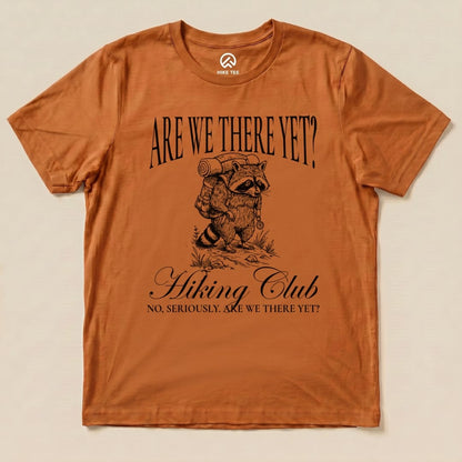 Graphic t-shirt in orange featuring a raccoon with a backpack and the slogan "Are We There Yet? Hiking Club" below it.