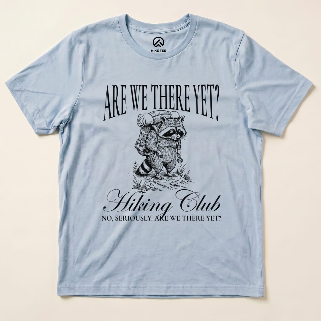 A light blue graphic t-shirt features a raccoon carrying a backpack with the slogan "Are We There Yet? Hiking Club.