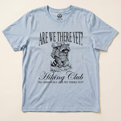 A light blue graphic t-shirt features a raccoon carrying a backpack with the slogan "Are We There Yet? Hiking Club.