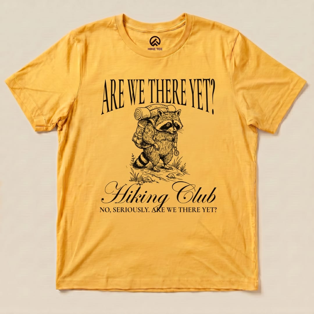 A yellow graphic t-shirt featuring a raccoon with a backpack and the phrase "Are We There Yet?" above "Hiking Club.
