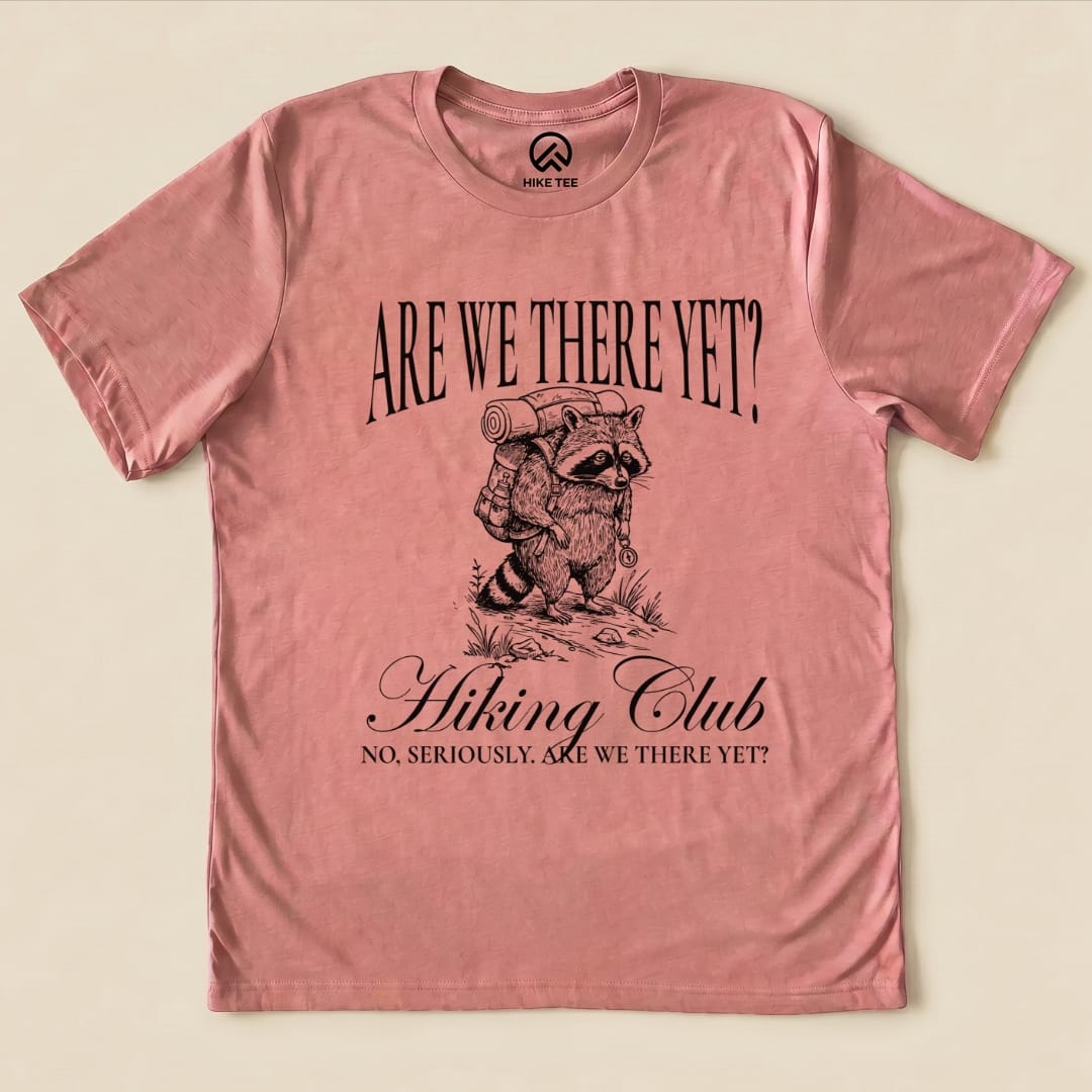 A pink graphic t-shirt features a raccoon with a backpack and the slogan, "Are We There Yet? Hiking Club," below.