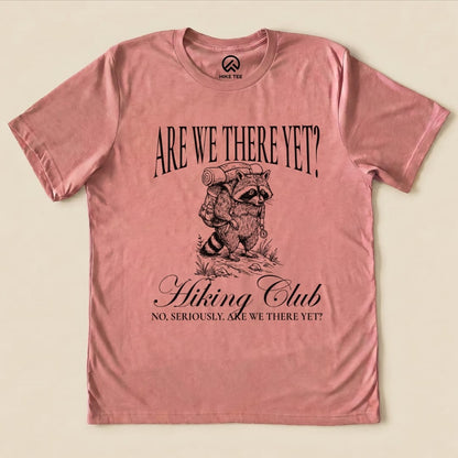 A pink graphic t-shirt features a raccoon with a backpack and the slogan, "Are We There Yet? Hiking Club," below.