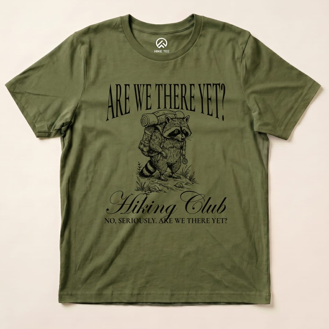A green graphic t-shirt featuring a raccoon carrying a backpack, with the text "Are We There Yet? Hiking Club" printed above.