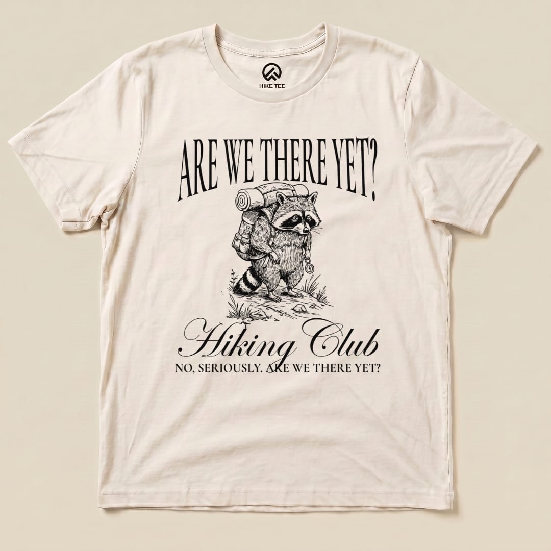 A graphic t-shirt featuring a raccoon with a backpack and the phrase "Are We There Yet? Hiking Club" printed above.