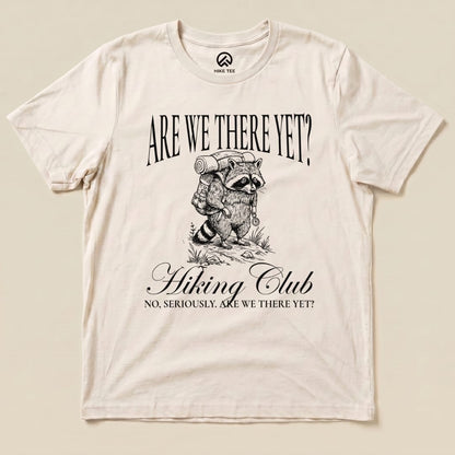 A graphic t-shirt featuring a raccoon with a backpack and the phrase "Are We There Yet? Hiking Club" printed above.