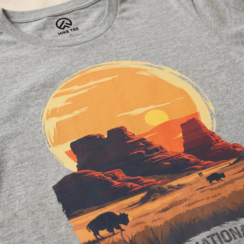 A gray graphic t-shirt featuring a sunset over Badlands National Park, with bison grazing in the foreground and bold text below.